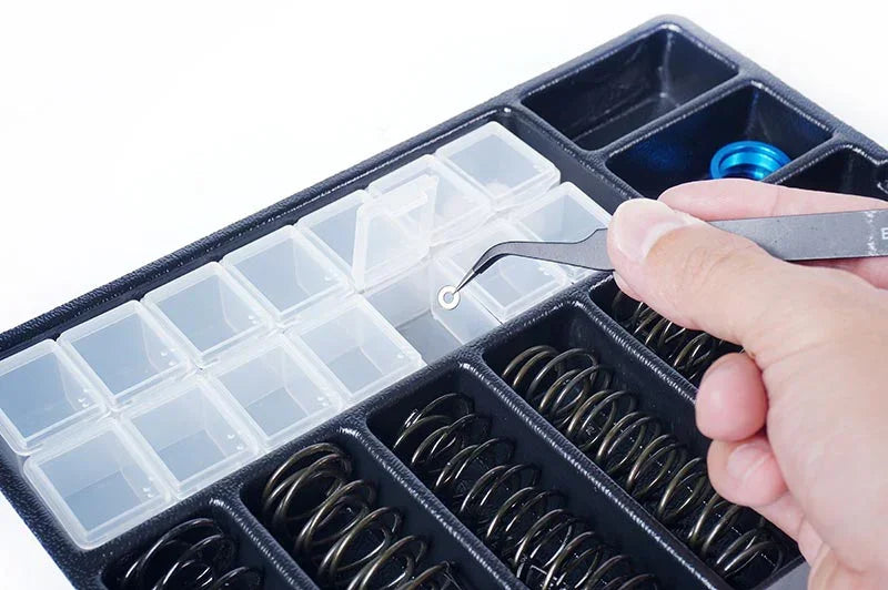 Hand using tweezers to pick a small RC part from organizer with springs and compartments