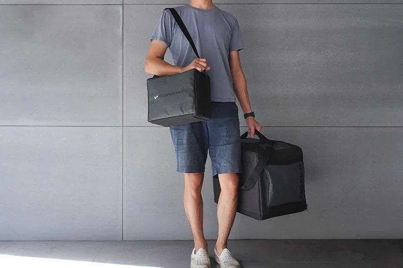 Person carrying two black RC pit bags with shoulder strap, standing against a gray wall